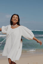 white Atoll dress standing on a beach with ocean waves in the background