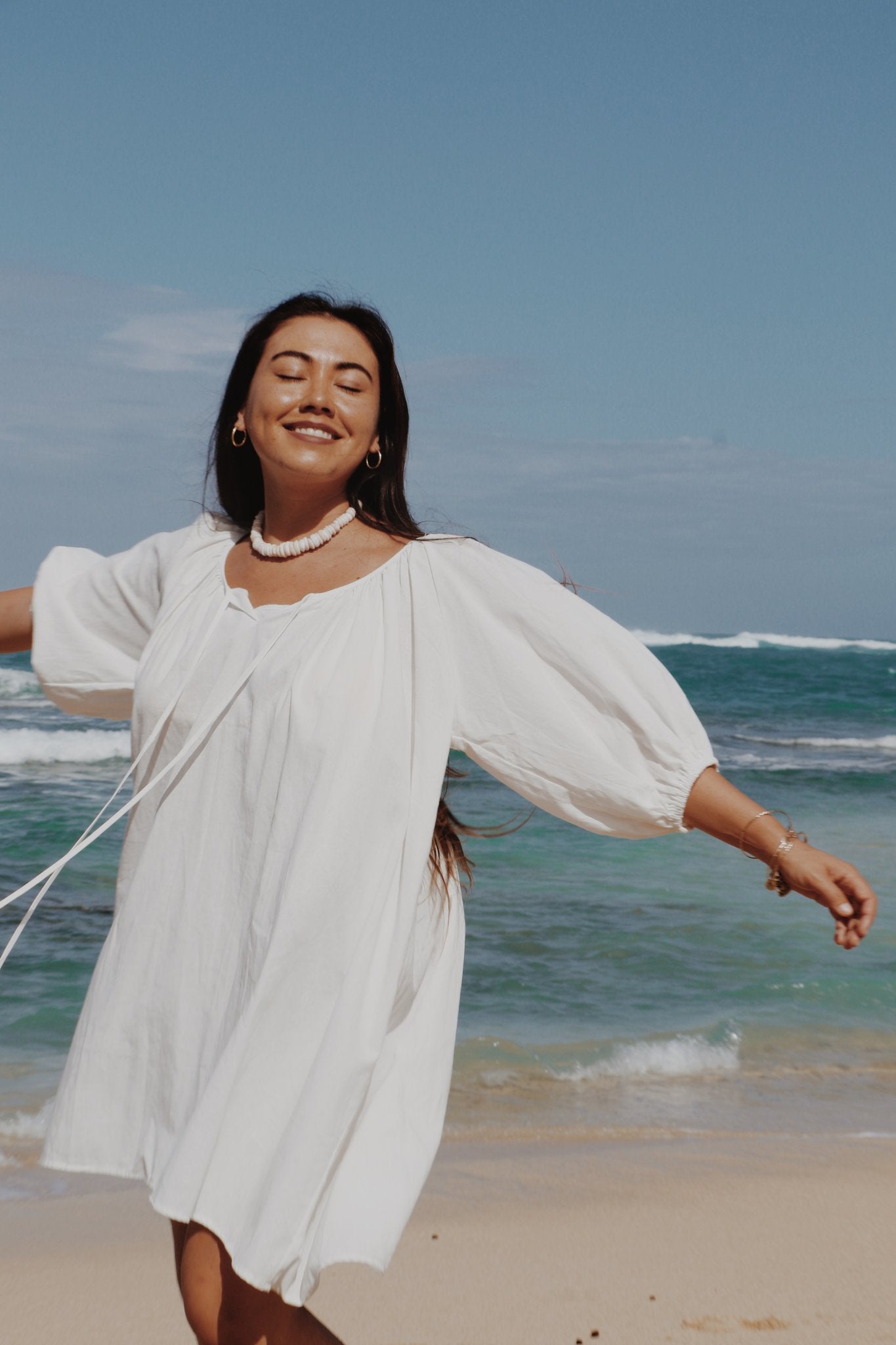 white Atoll dress standing on a beach with ocean waves in the background