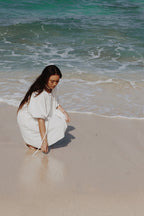 Woman in the Atoll white dress sitting on a beach with ocean waves in the background