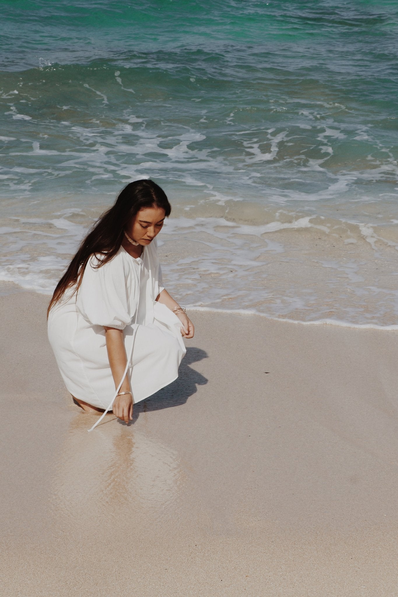 Woman in the Atoll white dress sitting on a beach with ocean waves in the background