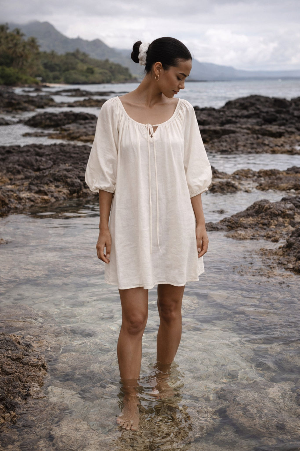 Woman in a Atoll white dress standing on a rocky beach with water and mountains in the background