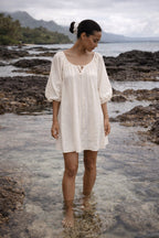 Woman in a Atoll white dress standing on a rocky beach with water and mountains in the background