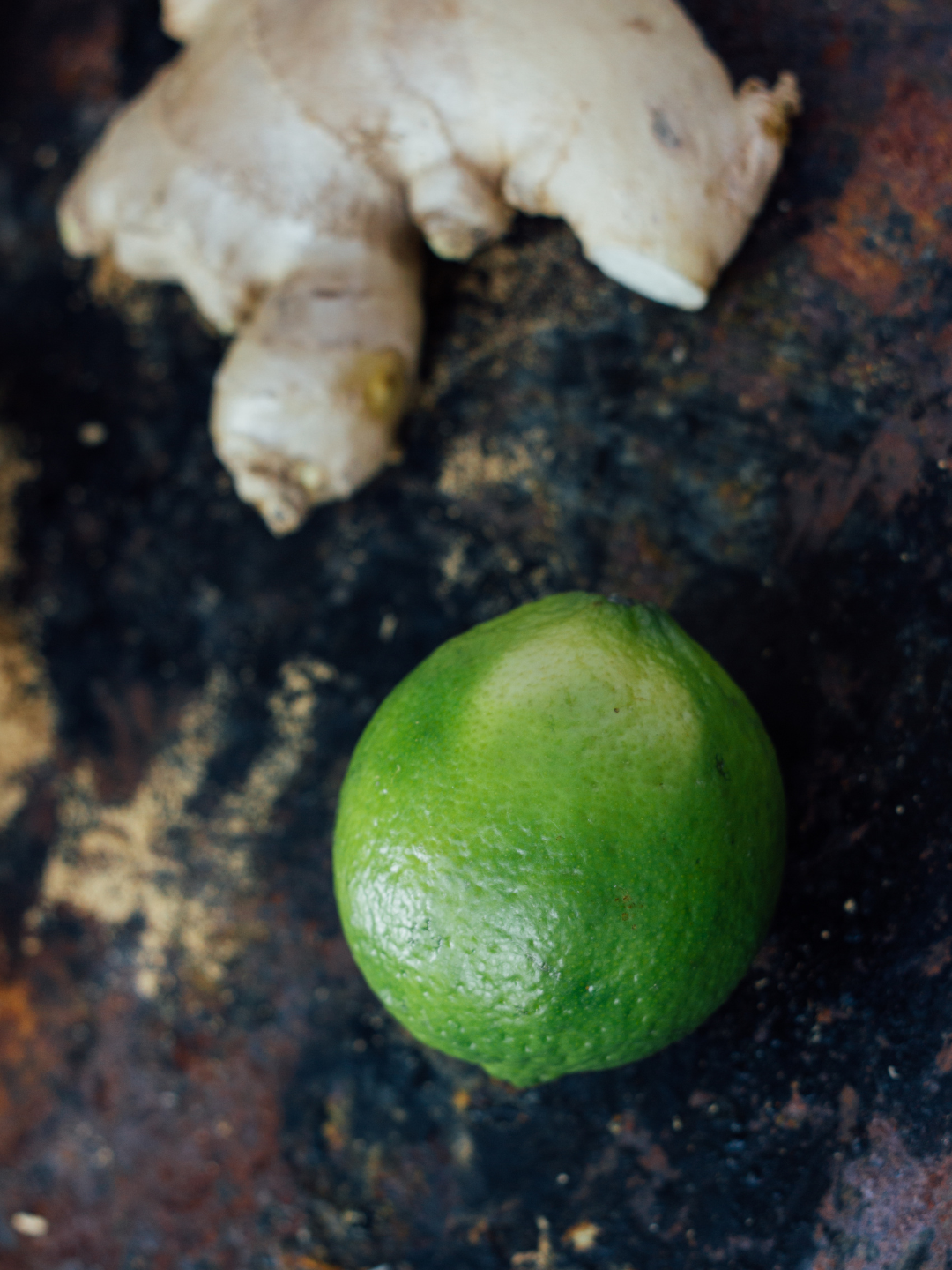 A whole green lime and a piece of ginger root resting on a dark, rustic, textured surface.
