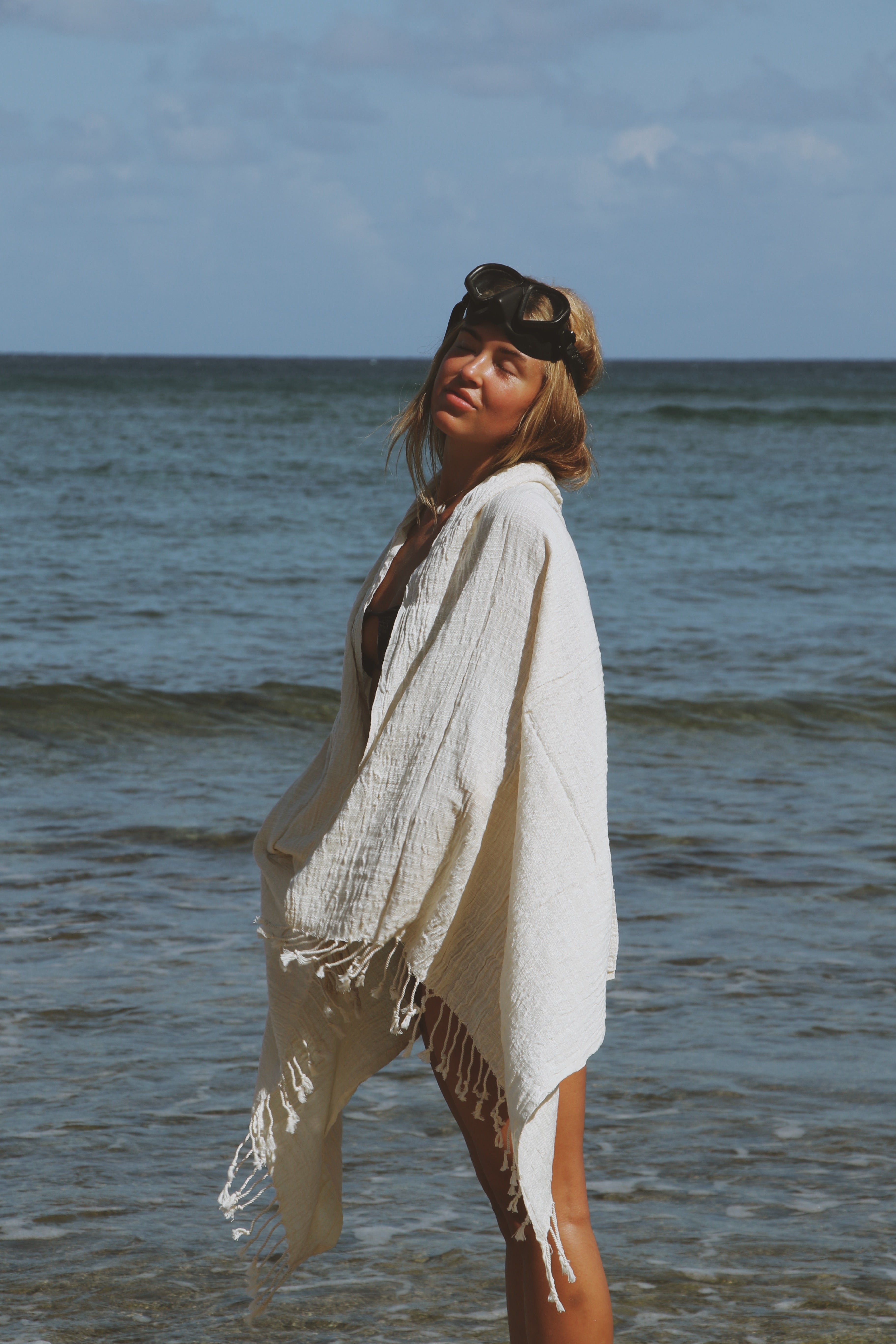 Woman standing on a beach with a white cover-up and sunglasses, ocean in the background