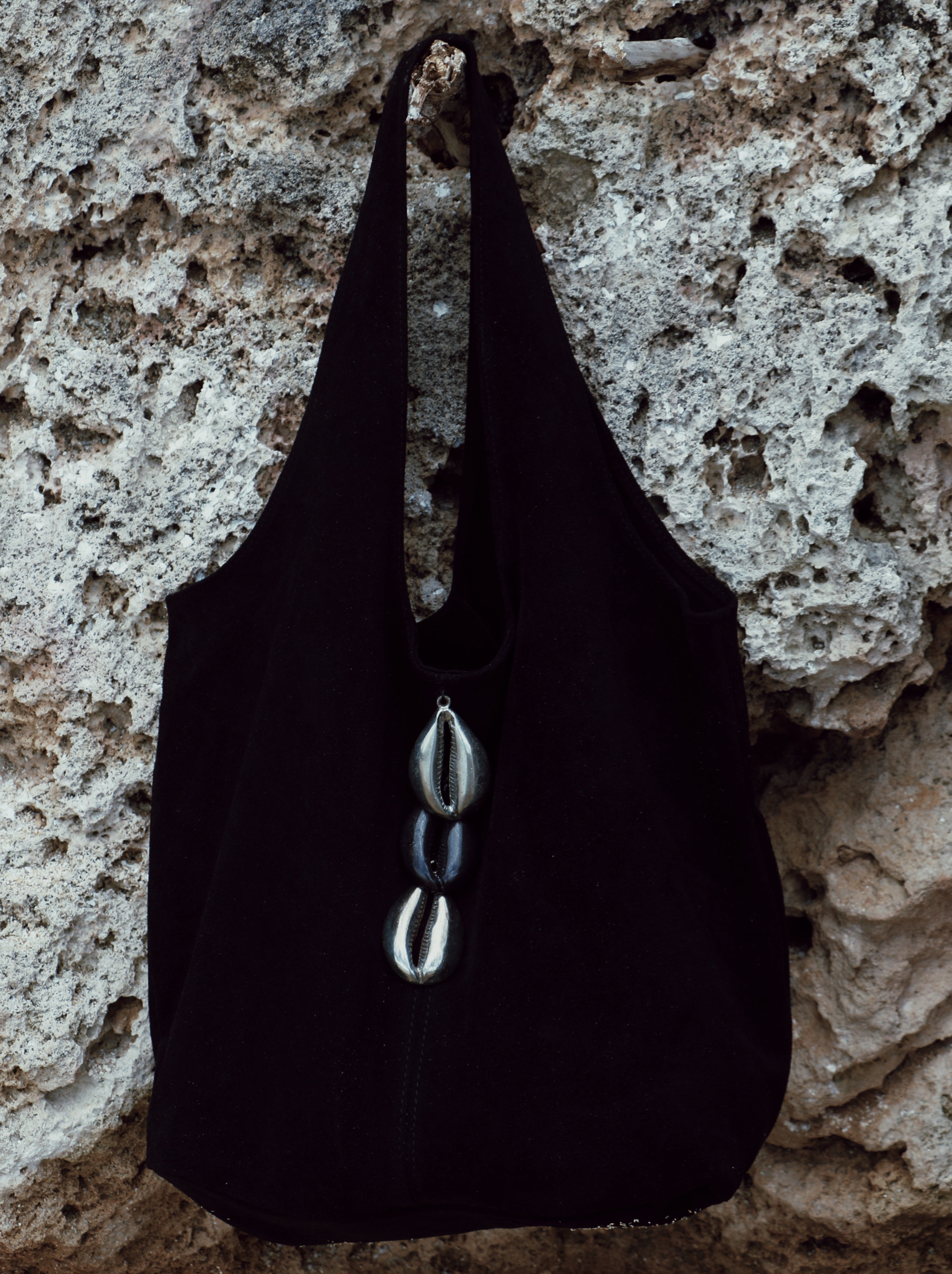 A black halter-style tote bag with three vertically stacked metallic cowrie shell charms, hanging against a textured light gray stone wall.