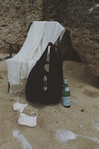 A black tote bag with silver-tone cowrie hardware stands on sandy ground next to a green bottle, with a white throw blanket draped over a chair and a pair of white flip-flops nearby. The scene is set against a rocky cliffside background.