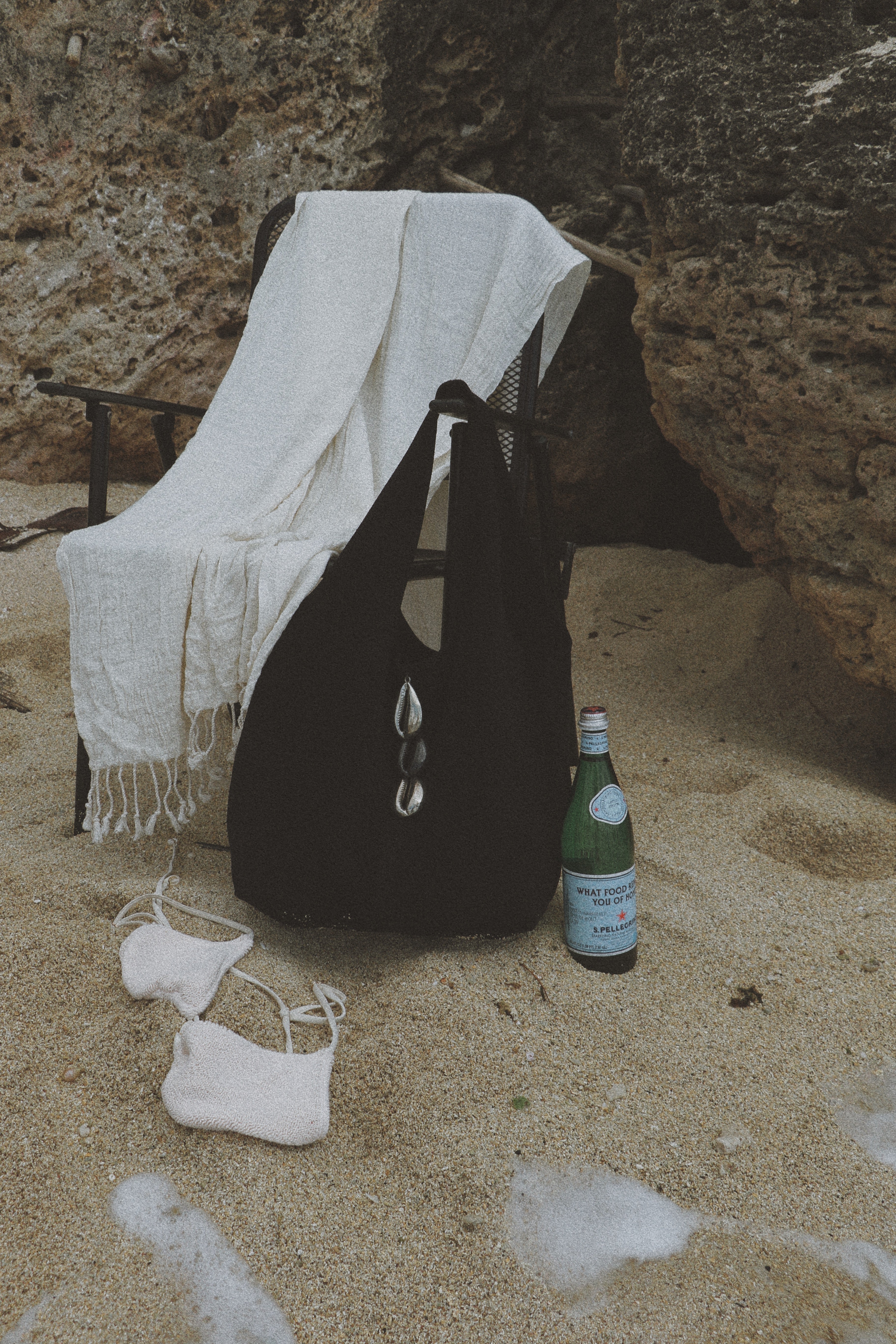 A black tote bag with silver-tone cowrie hardware stands on sandy ground next to a green bottle, with a white throw blanket draped over a chair and a pair of white flip-flops nearby. The scene is set against a rocky cliffside background.