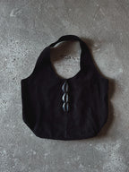 A black shoulder bag with a wide strap and three black cowrie accents down the center, laid flat on a textured gray concrete surface.