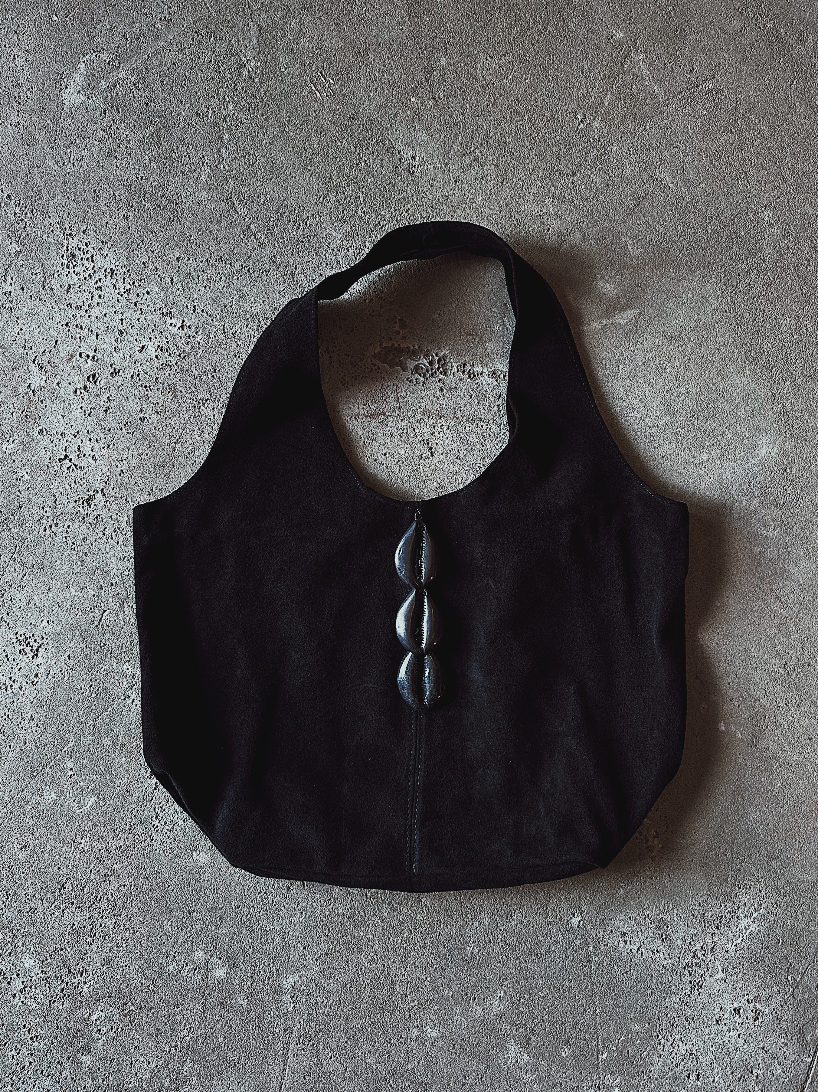 A black shoulder bag with a wide strap and three black cowrie accents down the center, laid flat on a textured gray concrete surface.