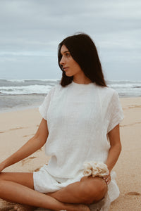 A woman dressed in white linen sits peacefully on the beach, holding a natural bath sponge as part of her ocean-inspired skincare ritual.