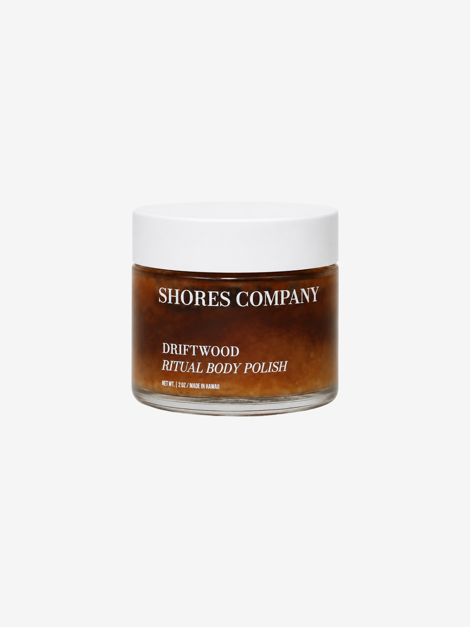 Shores Company Driftwood Ritual Body Polish in a clear glass jar with a white lid, showing the amber-brown polish inside, on a light gray background.