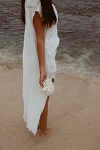 A woman in a white linen dress holding a natural bath sponge near the shore, embodying sustainable skincare.