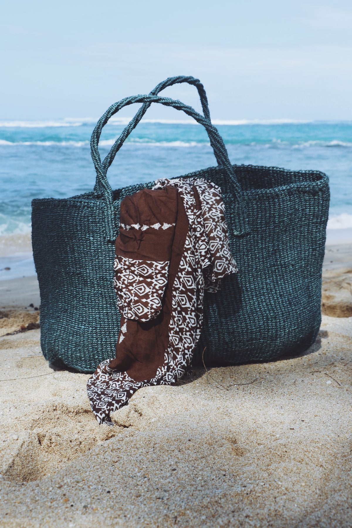 A large woven tote bag sits on sandy beach ground with the ocean and sky in the background. A brown and white patterned scarf is draped over the front of the bag.