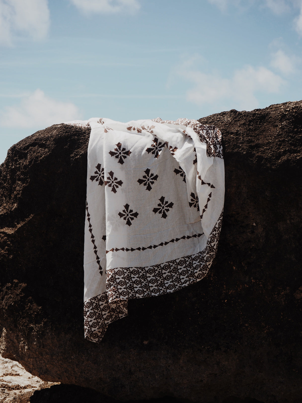 A white cotton bandana or scarf with black floral and geometric embroidery, draped over a large dark rock under a blue sky.