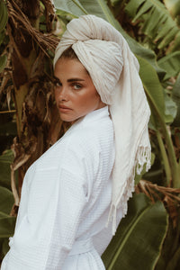 A woman in the Noosa Bathrobe, exuding tropical luxury, with towel wrapped on head, framed by vibrant banana leaves.
