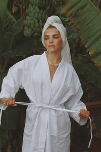 Model adjusting the waist tie of the Noosa Bathrobe, made of soft waffle-textured cotton, with lush greenery in the background.