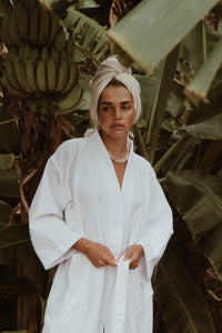 A woman wearing the Noosa Bathrobe in white cotton waffle fabric, standing in a tropical setting with banana leaves.