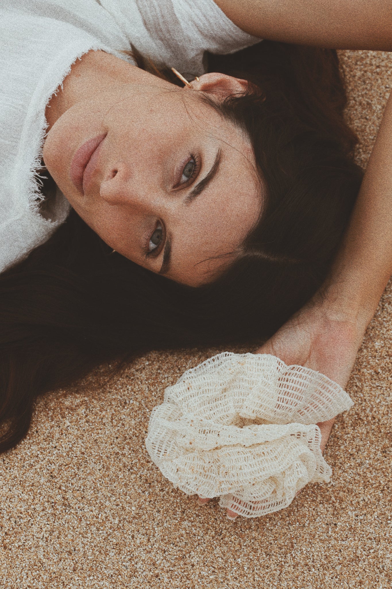 A woman lying on soft sand, holding an organic bath sponge, embracing natural self-care.