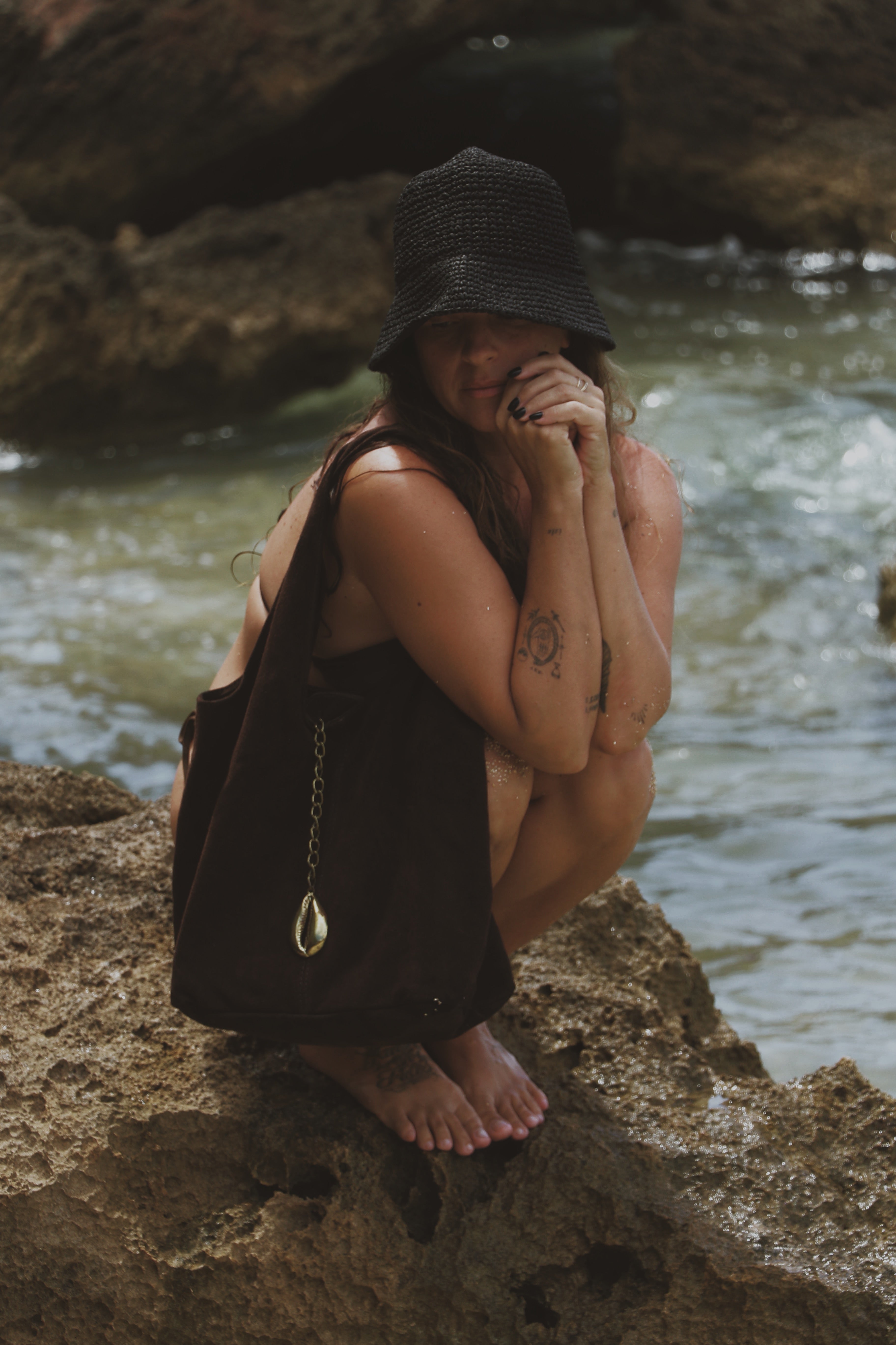 A woman crouches on a rocky shoreline with water in the background, wearing a black woven bucket hat, a brown bikini, and carrying a black shoulder bag with a gold cowrie charm.