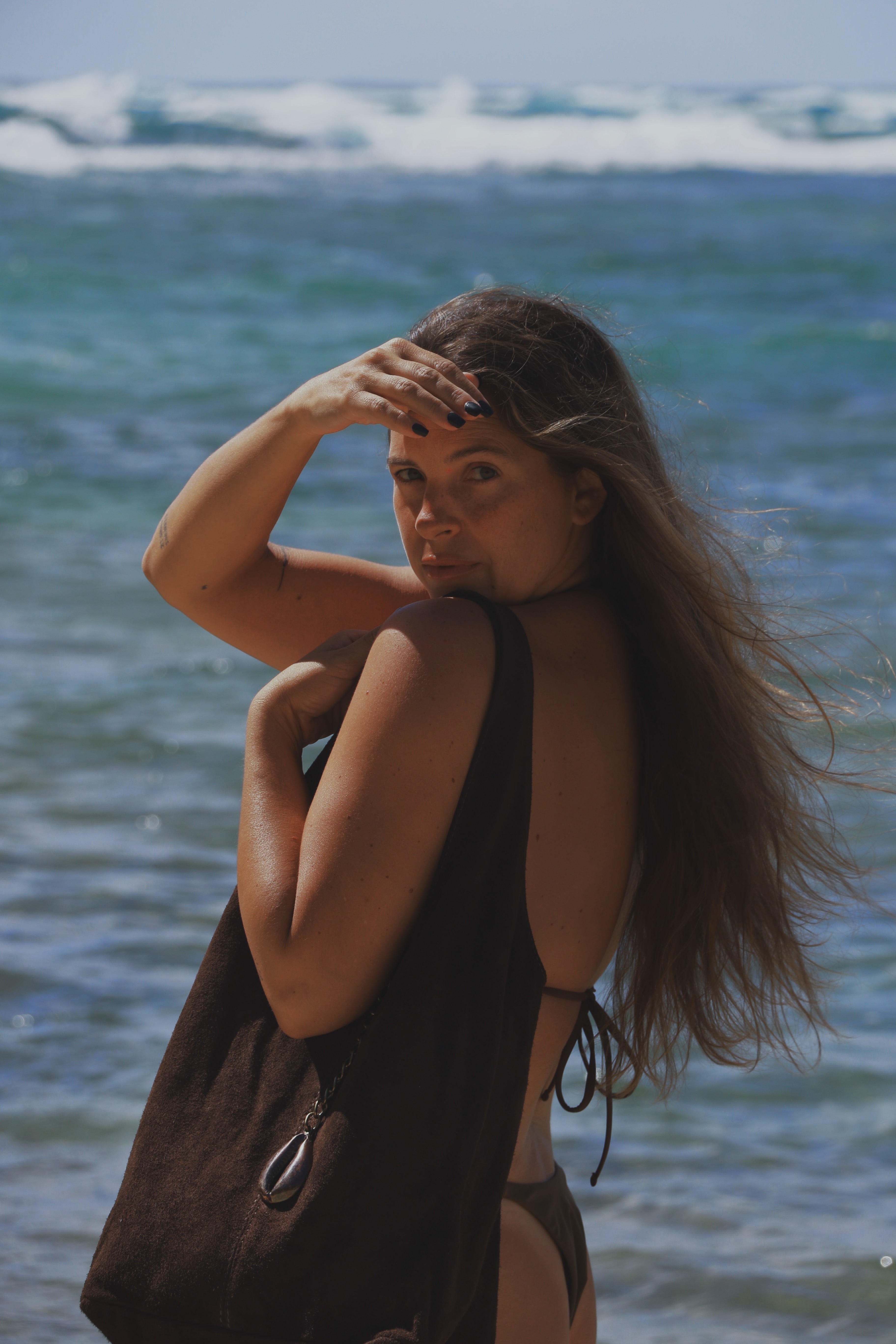 A woman with long brown hair stands on a beach in front of the ocean waves, wearing a dark brown bikini and carrying a matching dark brown tote bag.