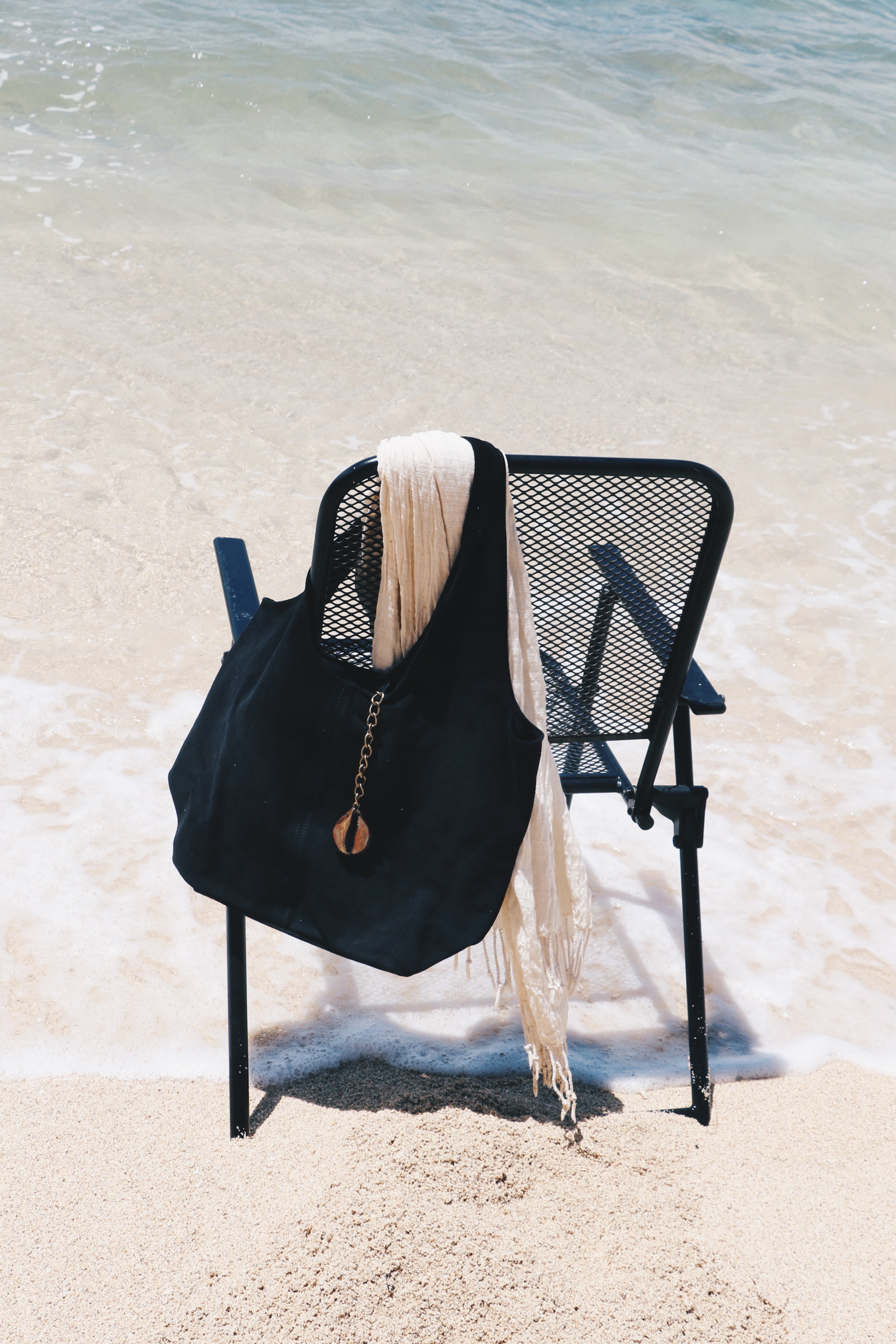 A black suede tote bag with a gold-toned cowrie charm and chain strap, draped over a black metal beach chair on a sandy beach with ocean water in the background.