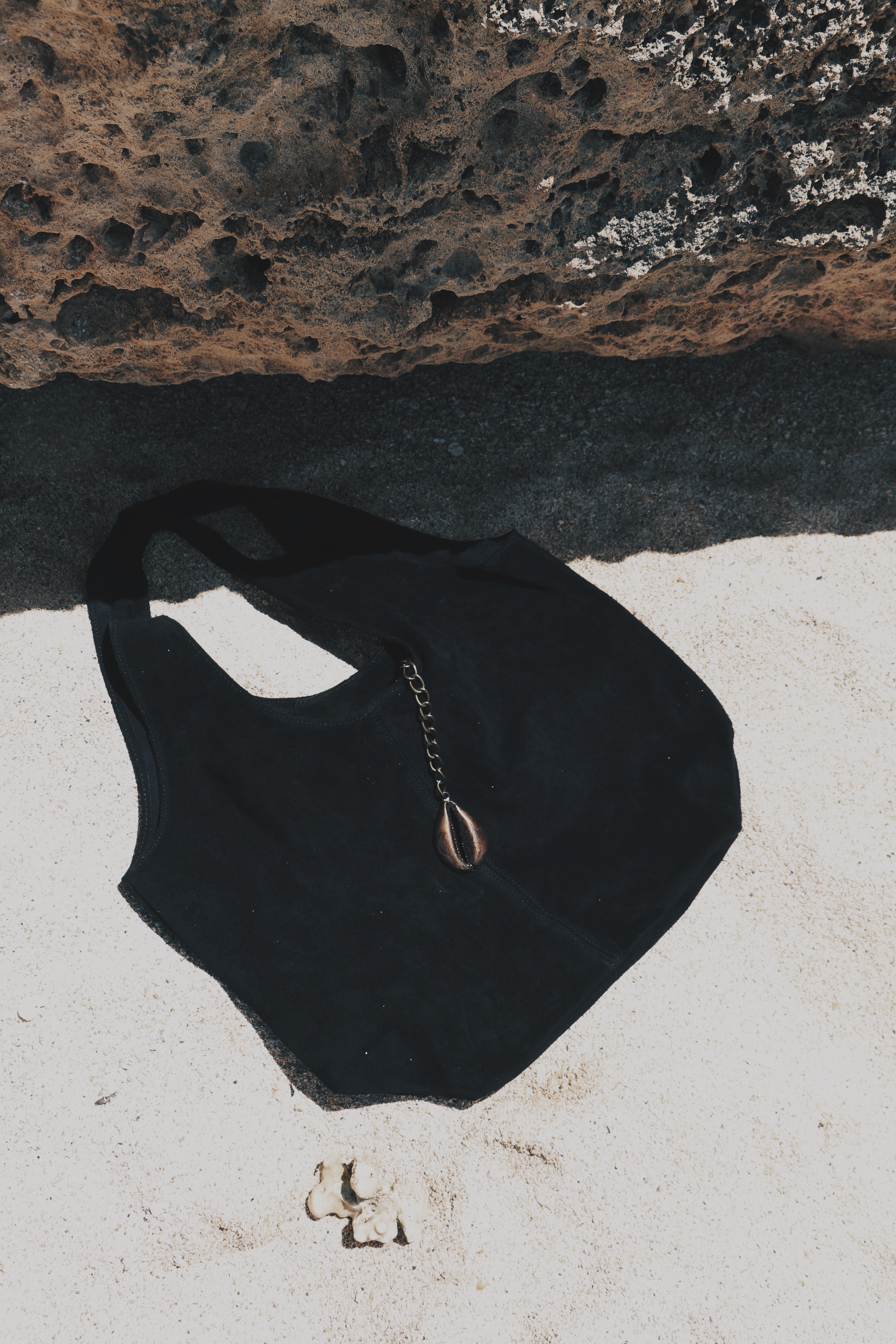 A black fabric shoulder bag with a gold chain and teardrop cowrie pendant, laid flat on a white surface with a dark textured rock background.