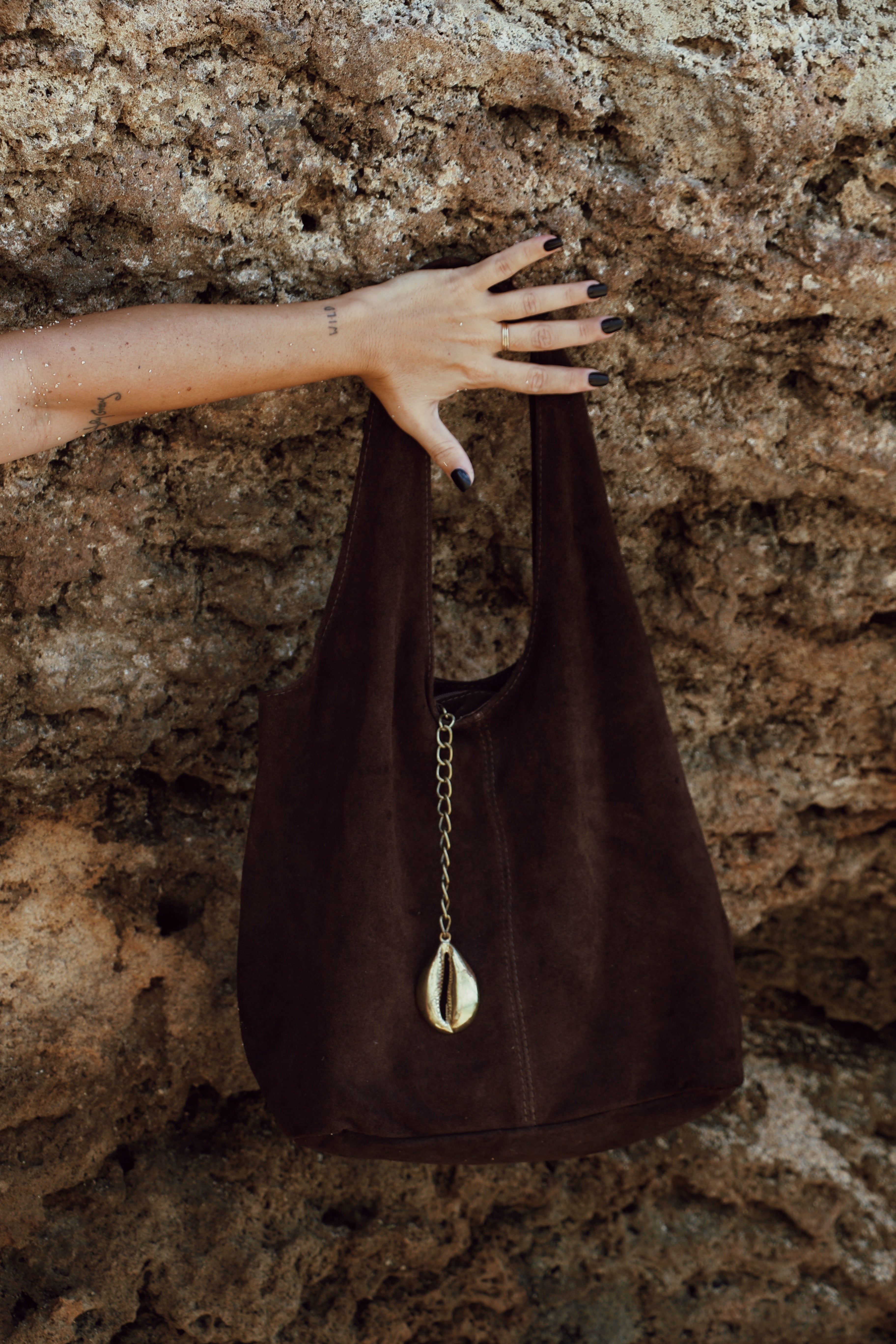 A dark brown suede hobo-style shoulder bag with a gold chain and teardrop cowrie pendant, held by a person's hand against a textured rock wall.