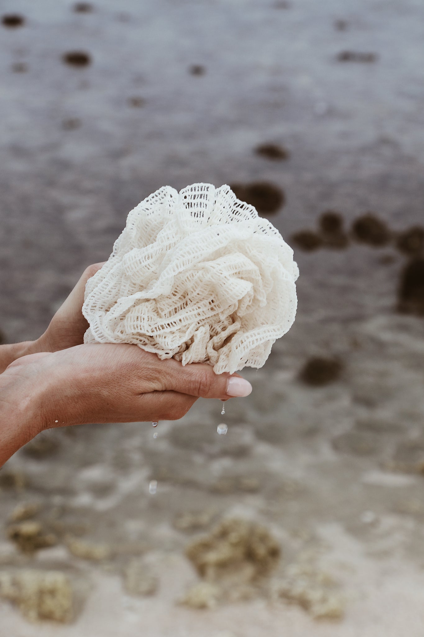 A close-up of a hand holding an organic bath sponge, dripping water against the ocean backdrop.