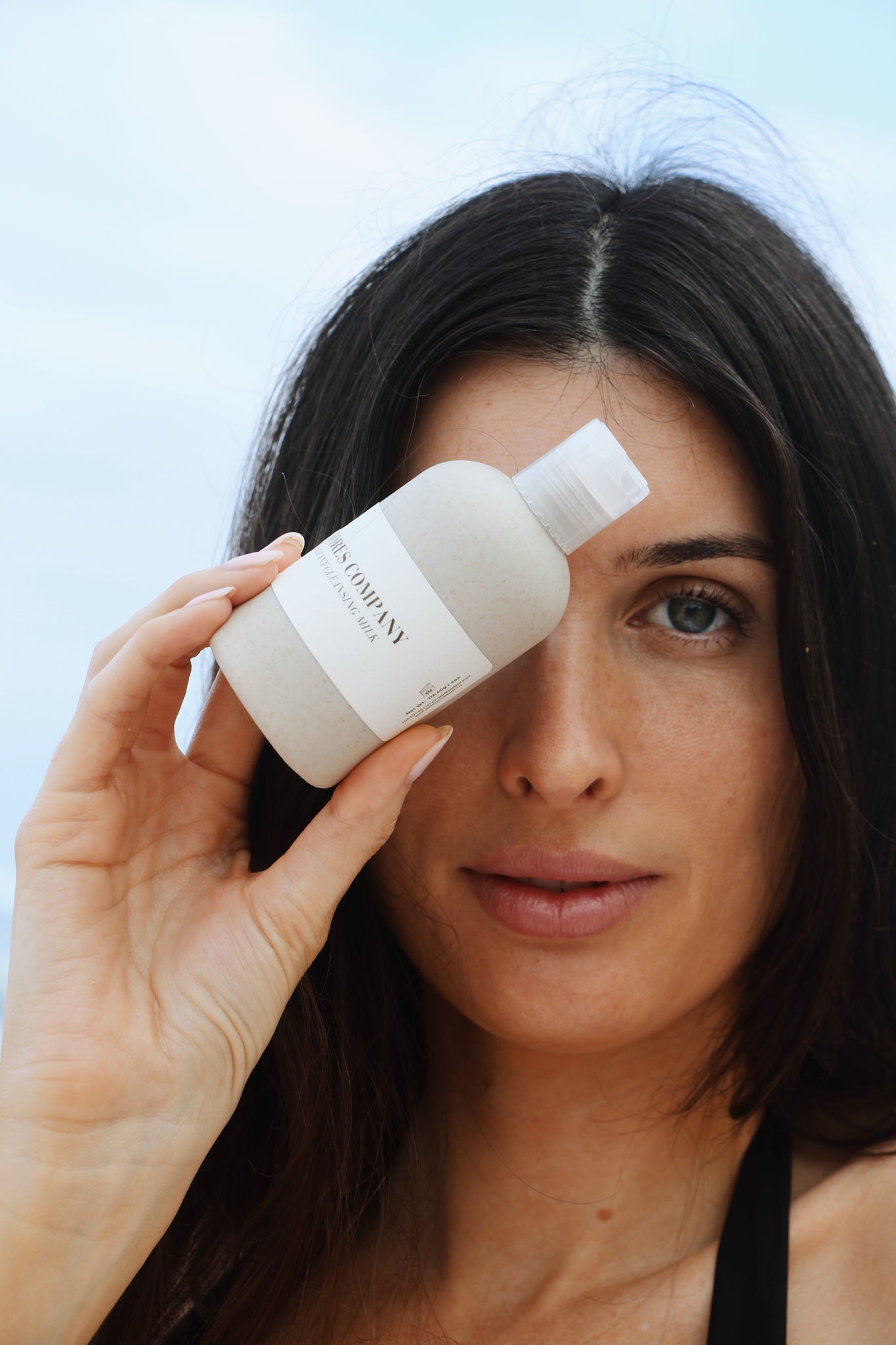 A woman holding Shores Company Radiant Cleansing Milk against a blue ocean backdrop, embodying the essence of sustainable, Hawaii-made skincare.