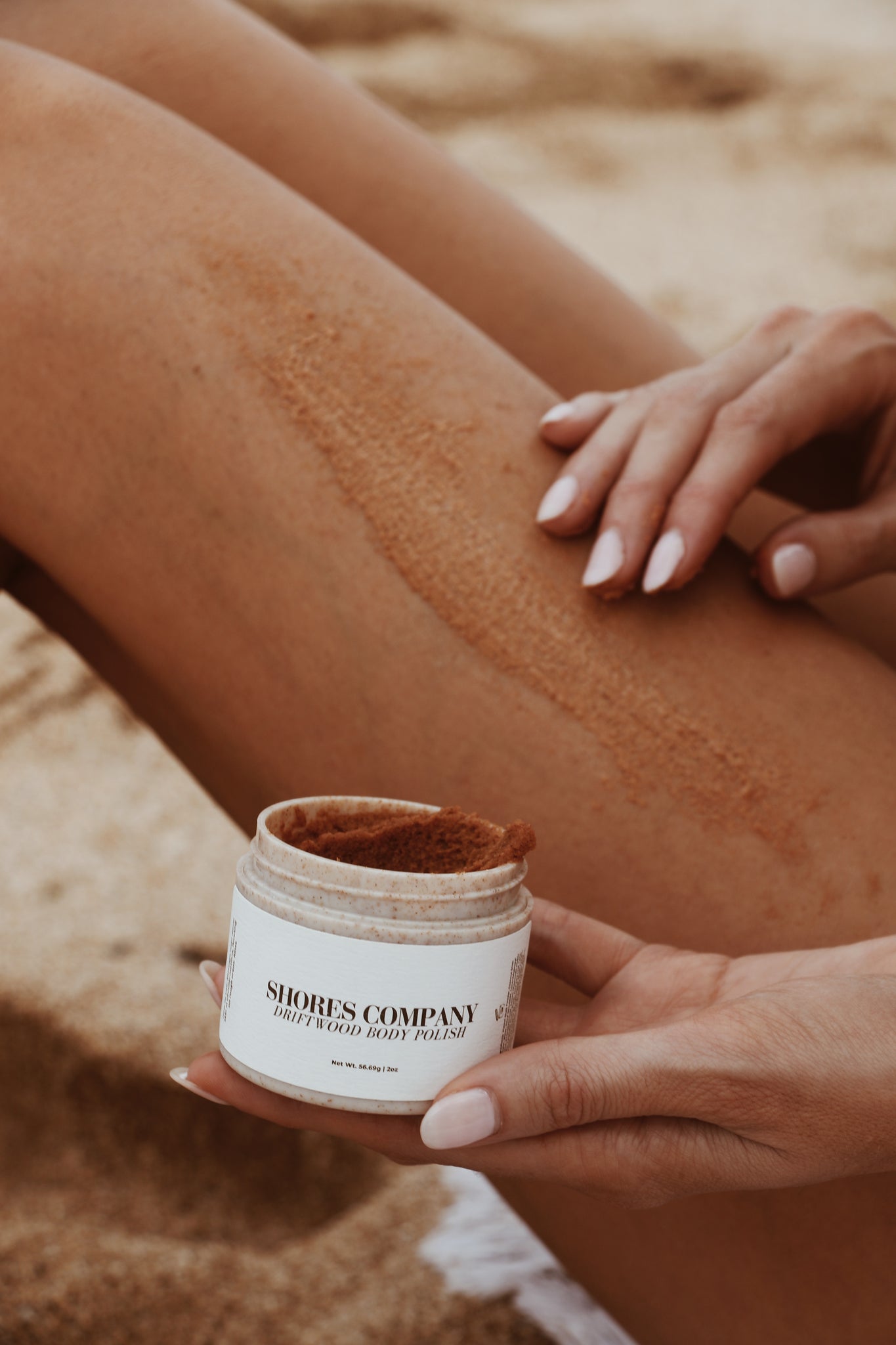  A model showcasing the smooth exfoliation of Shores Company’s natural Driftwood Body Polish on her leg, perfect for beach-ready skin.