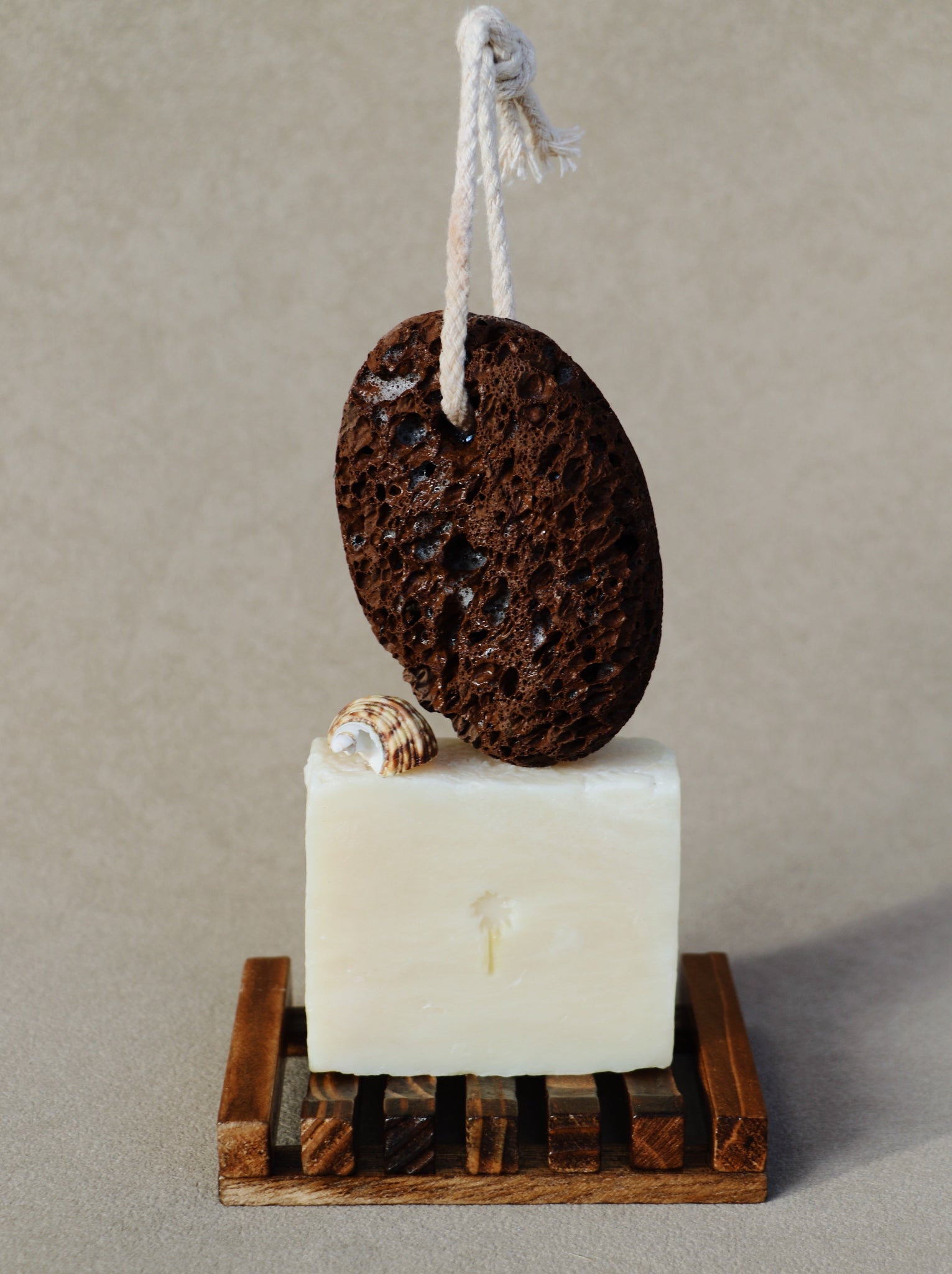 A handmade bath product set featuring a dark brown porous lava stone hanging from a white rope, resting on top of a rectangular bar of off-white soap, which sits on a small wooden slotted tray. A small seashell is tucked into the side of the soap.