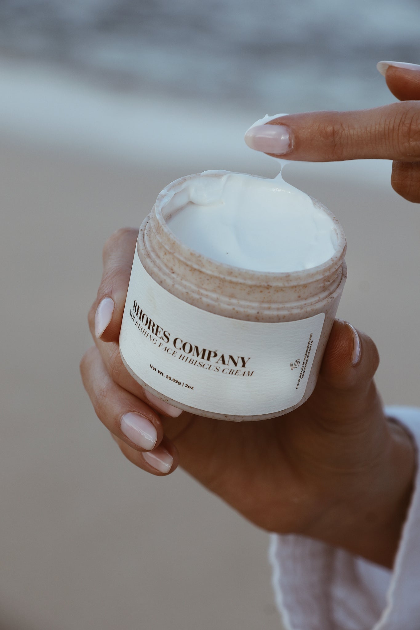 A hand dipping into Shores Company Nourishing Face Hibiscus Cream, demonstrating its luxurious hydration and natural formula.