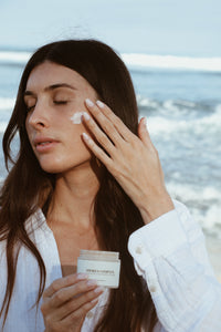 A woman applying Shores Company Nourishing Face Hibiscus Cream by the ocean, highlighting natural skincare from Haleiwa, Hawaii.