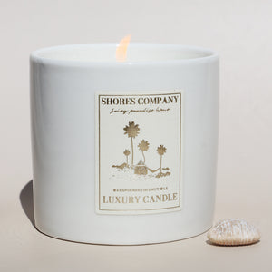 Shores Company luxury coconut wax candle in a white ceramic vessel, softly glowing with a warm flame.