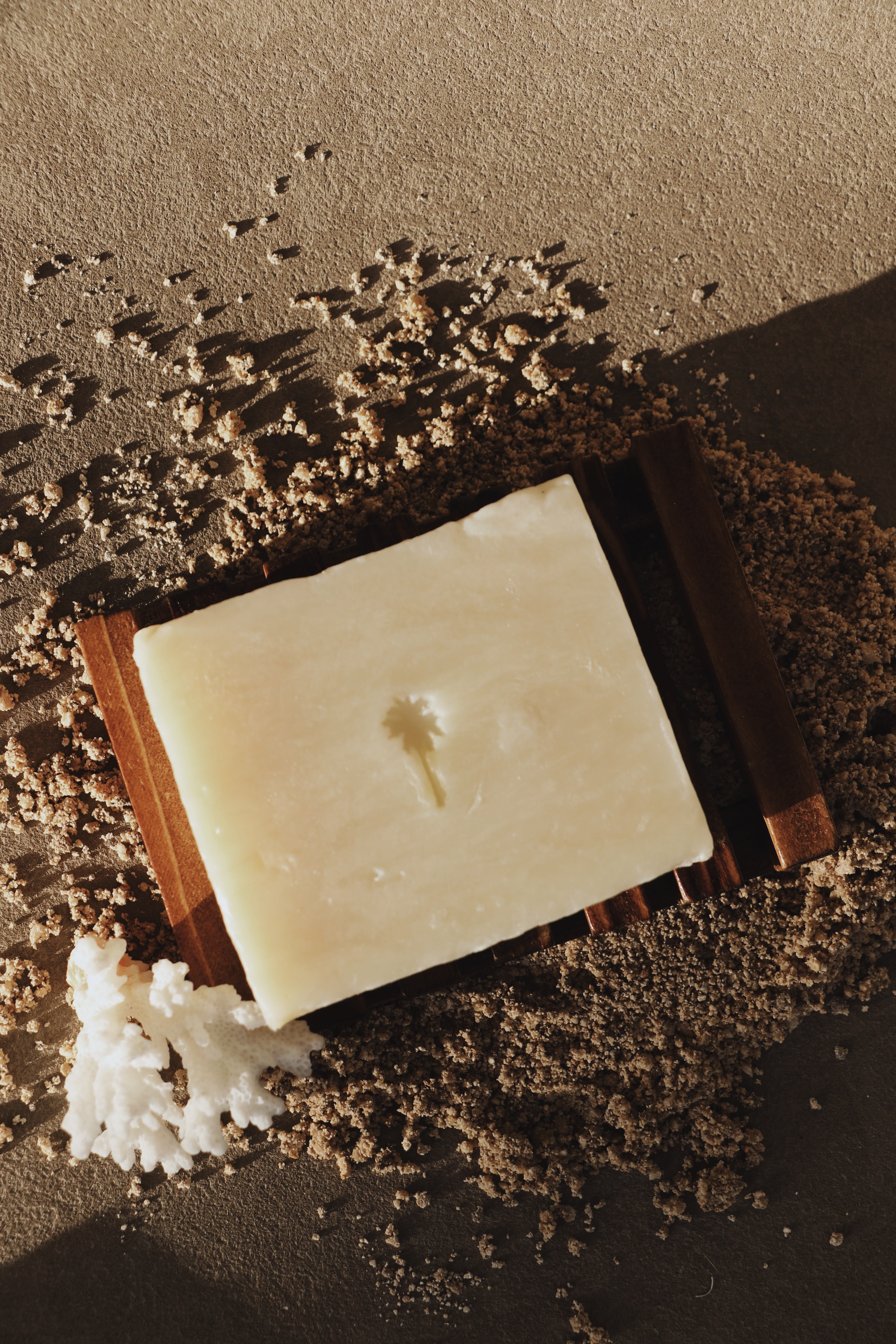 A rectangular bar of pale cream soap resting on a dark wooden slotted board, surrounded by coarse brown sand and a small white coral piece, in warm directional light.