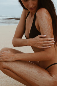 A model sitting on the sand, rubbing Shores Company Driftwood Body Polish on her arm and leg for a natural beachside skincare ritual.