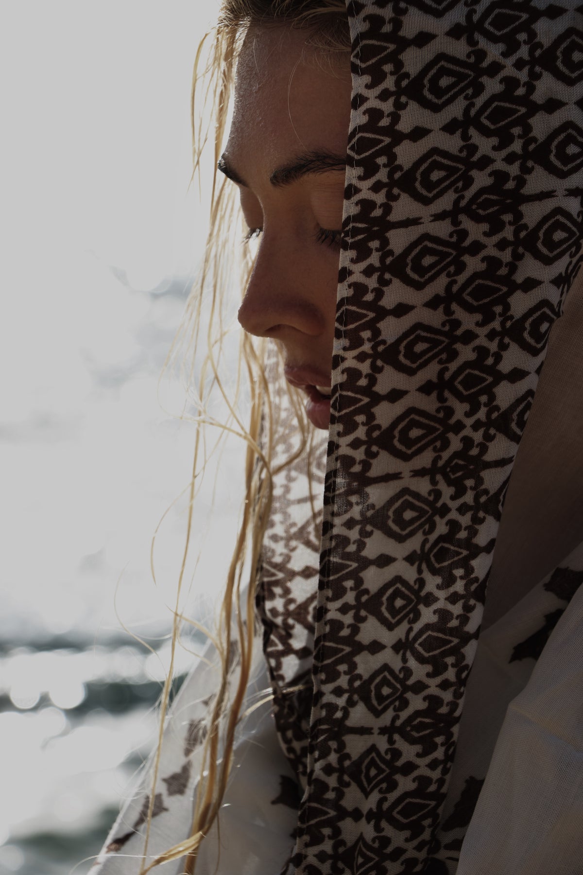 A close-up lifestyle shot of a woman wearing a white hood or scarf with a black geometric tribal pattern, partially covering her face against a bright, overexposed outdoor background.